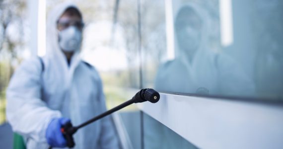 Person in white chemical protection suit doing disinfection of public areas to stop spreading highly contagious corona virus.